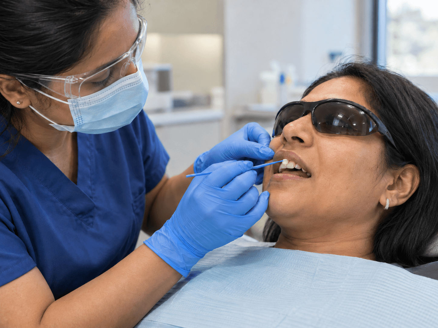 A hygienist wearing gloves, a mask and protective glasses applies varnish to a patient's upper front tooth.