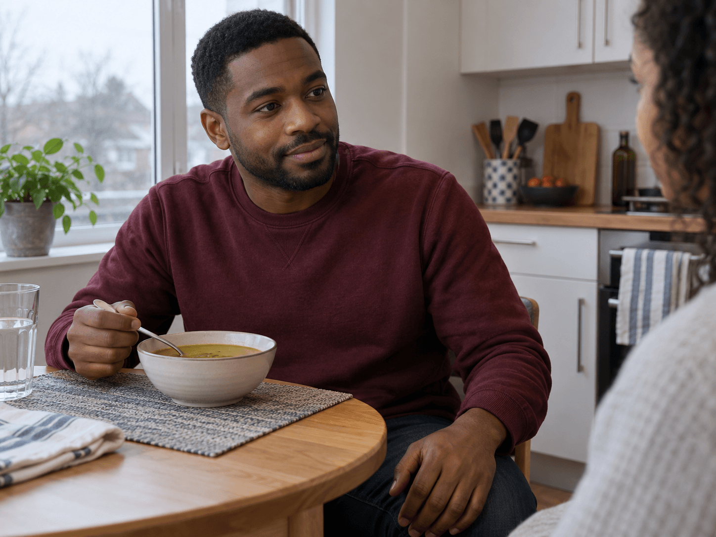 A man eats a bowl of soup while talking with his partner at home.