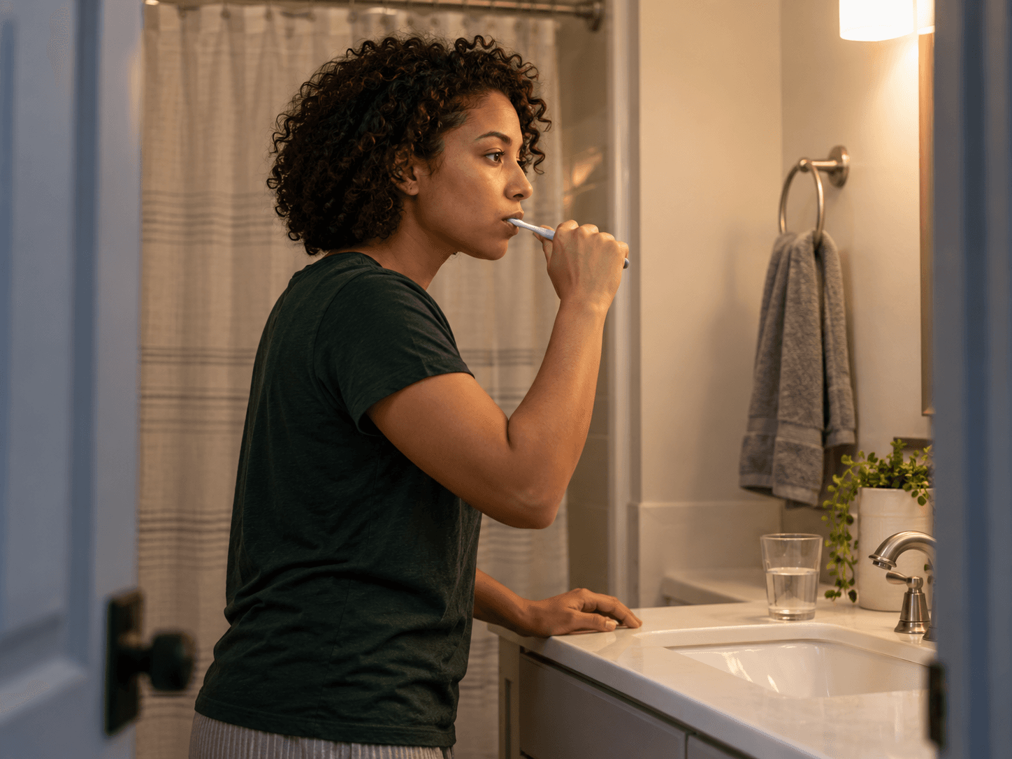 A woman brushes her teeth at her bathroom sink in the evening.